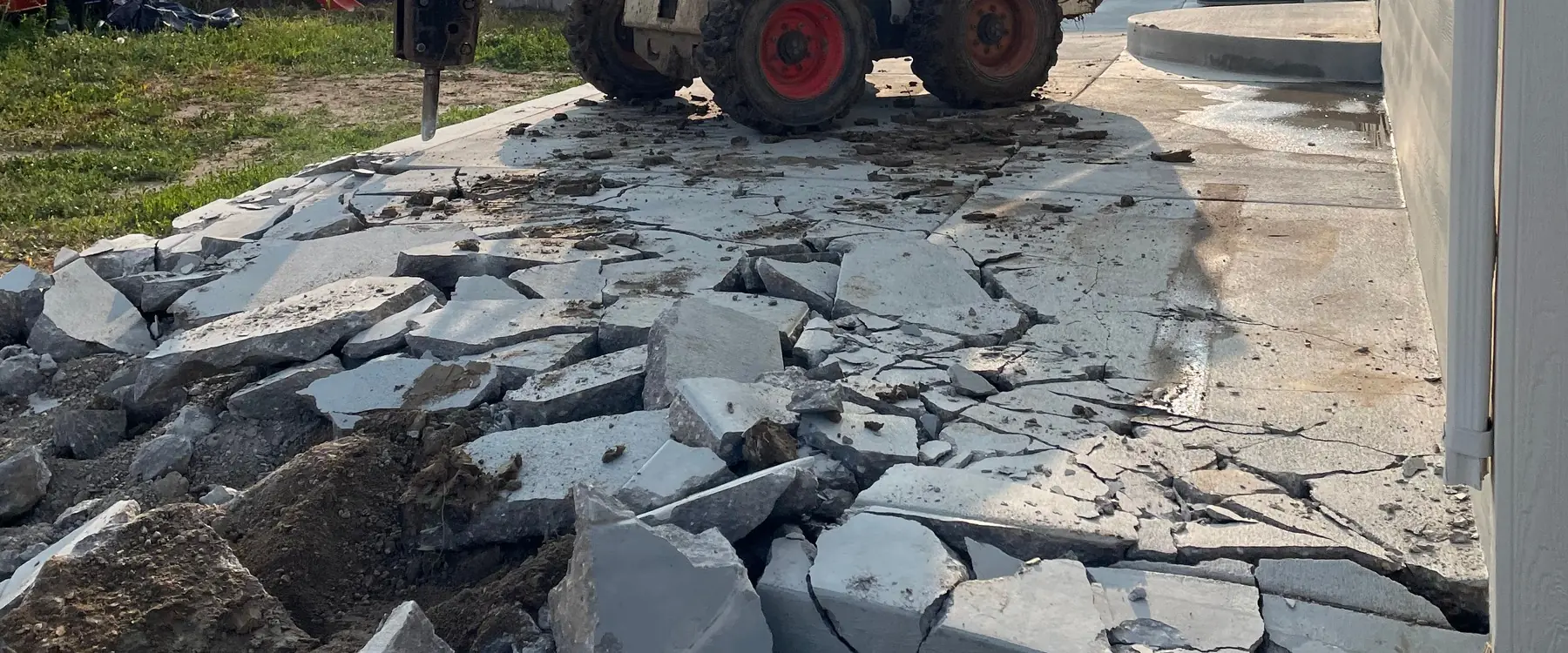Old concrete broken out beside a house, with a skid steer in the background
